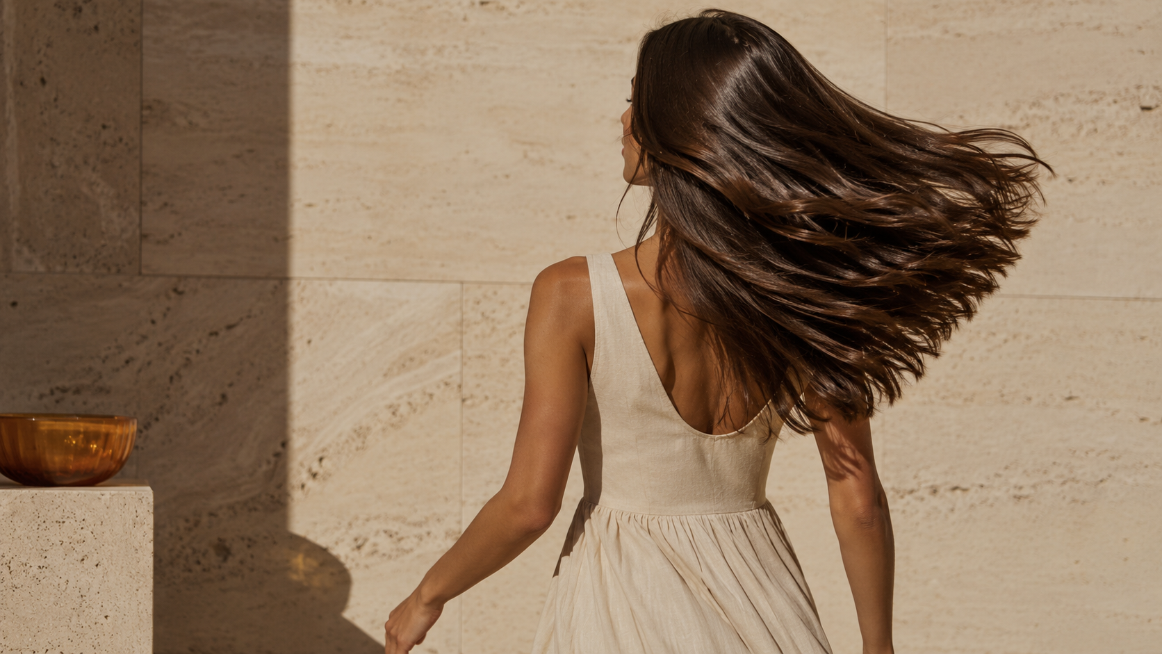 Cabelo castanho em movimento com reflexo intenso, sob luz quente, diante de parede de travertino